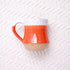 A hand-thrown ceramic mug with a narrow lip and thumb rest on the handle, featuring a Coral Conch colorway with orange and beige tones.